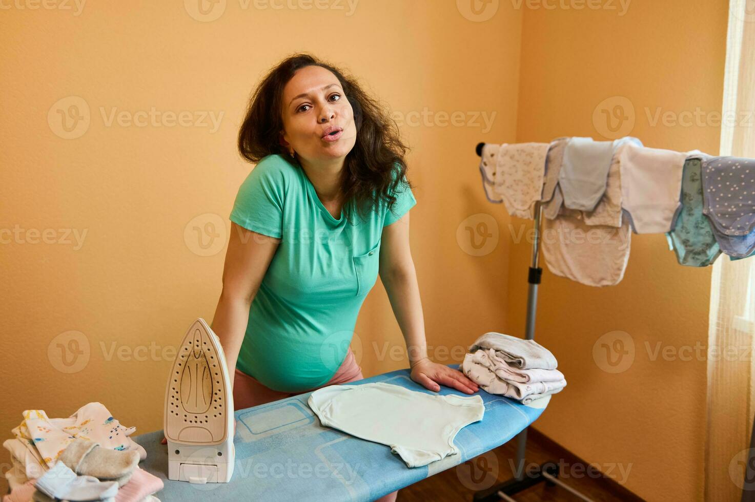 uitgeput zwanger vrouw in 3d trimester van zwangerschap, staand door strijken bord, stomen pasgeboren baby kleren foto