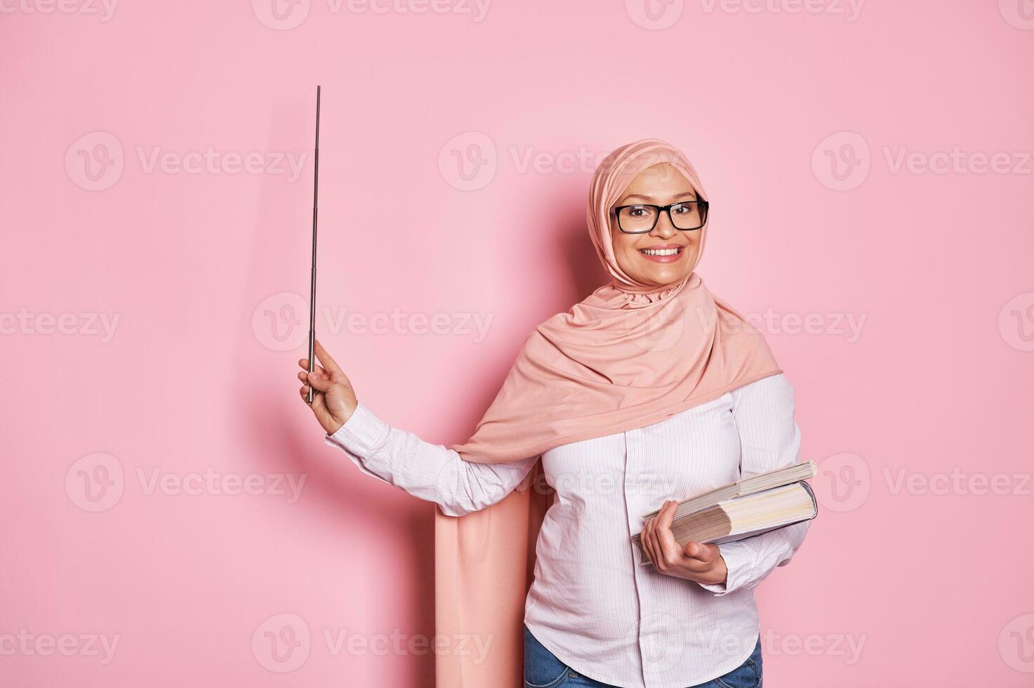 vrolijk ervaren midden- oud Arabisch musim leraar in roze hijaab, points met wijzer Bij kopiëren ruimte Aan roze achtergrond foto