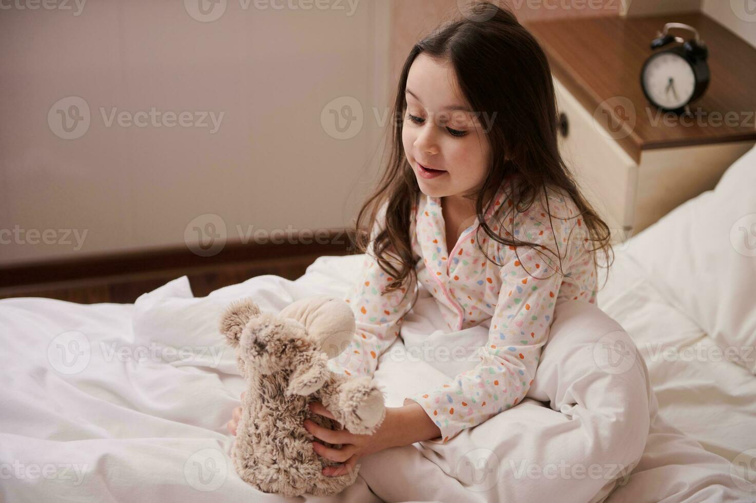 aanbiddelijk weinig kind meisje in elegant pyjama, zittend Aan bed en spelen met haar pluche speelgoed- schapen voordat gaan naar bed foto