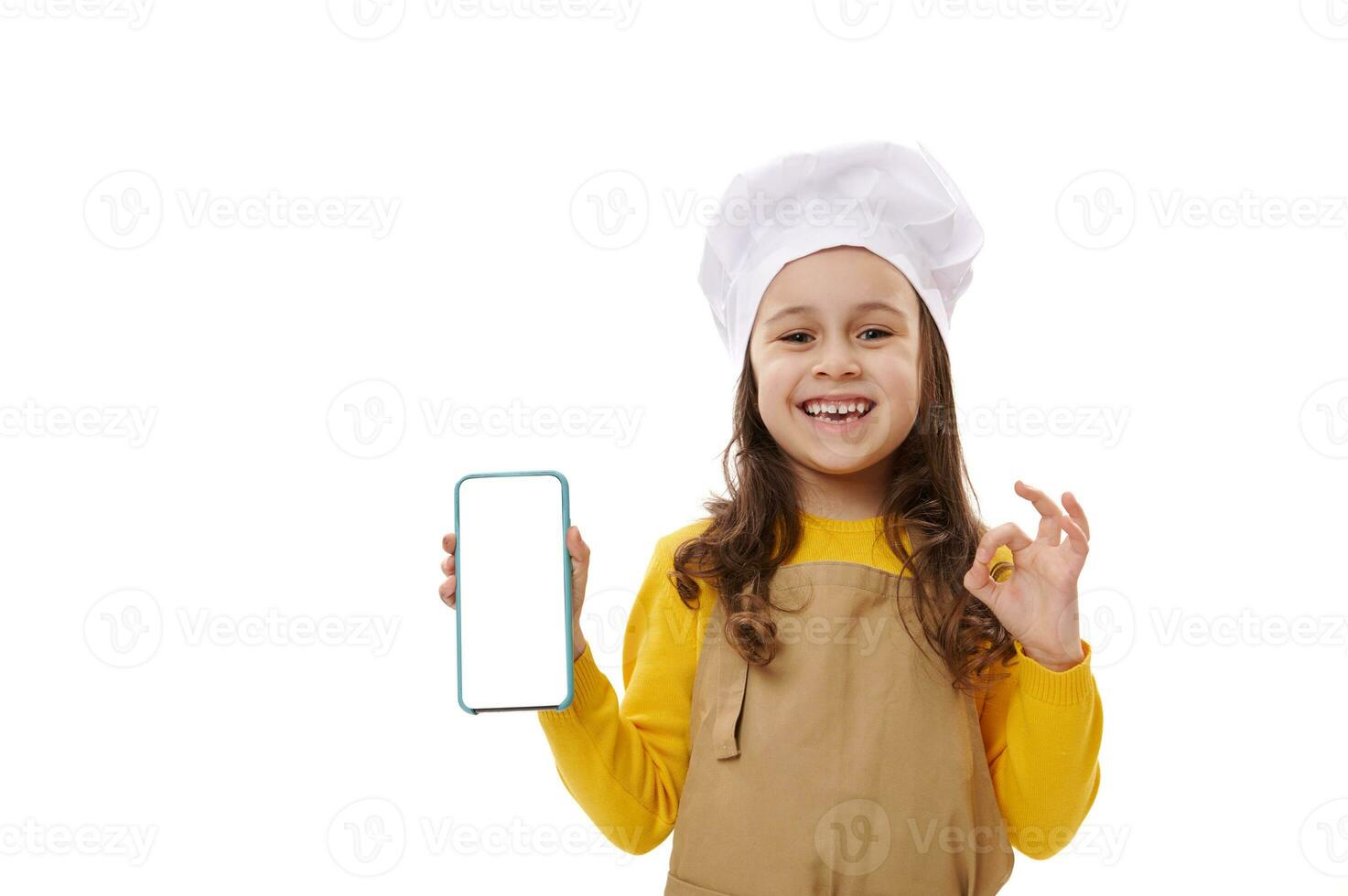 positief weinig kind meisje in chef hoed en schort, tonen smartphone met blanco wit scherm. kopiëren ruimte voor mobiel apps foto