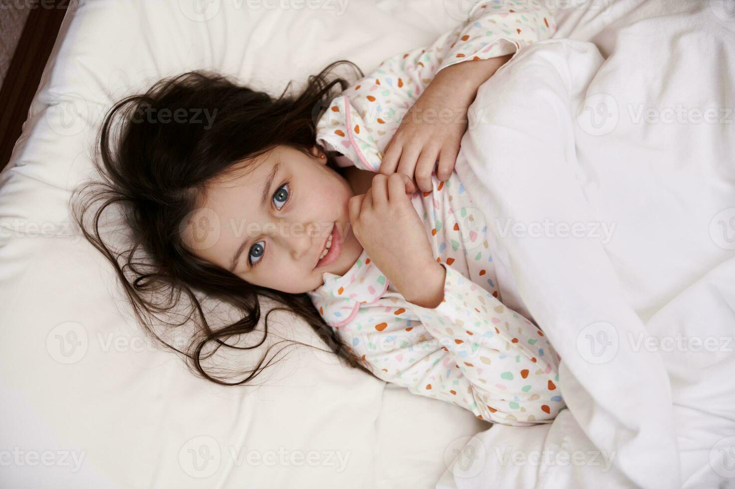 visie van bovenstaand van aanbiddelijk weinig meisje in pyjama, verpakt in wit knus donsdeken, aan het liegen Aan bed, op zoek Bij camera foto