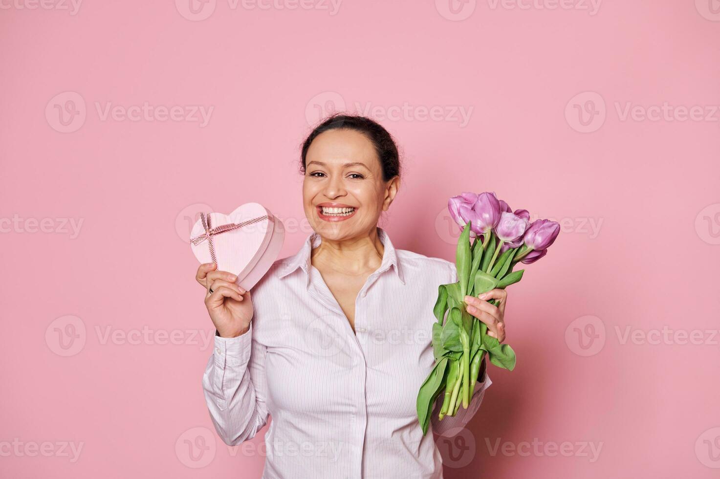 positief vrouw met boeket van Purper tulpen en geschenk doos, glimlachen een toothy glimlach, op zoek Bij camera, geïsoleerd Aan roze foto