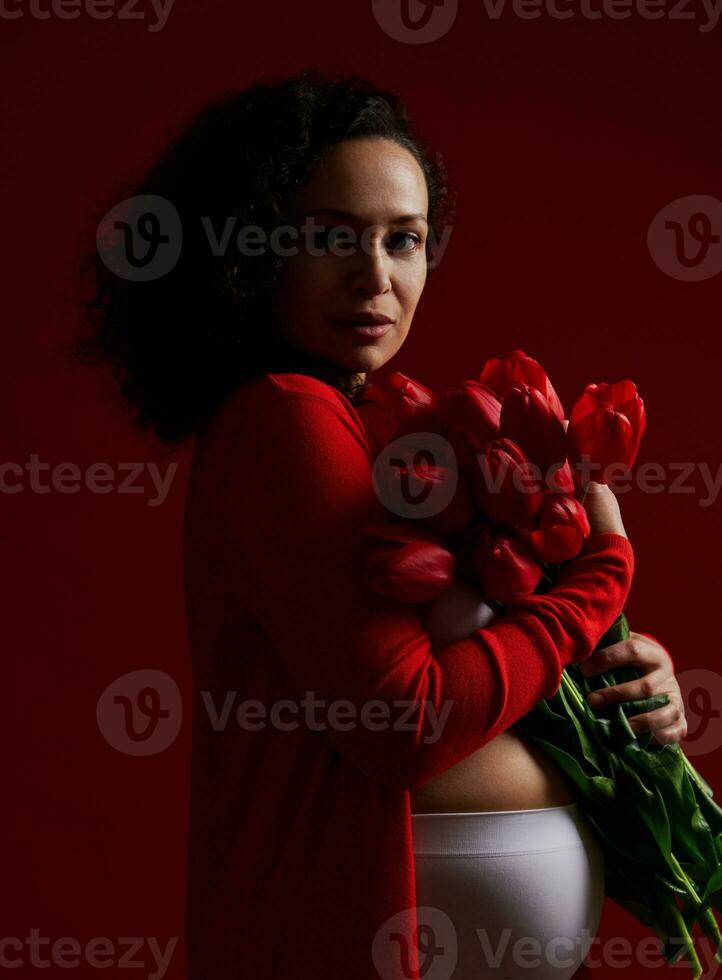 authentiek volwassen zwanger vrouw Holding een boeket van tulpen, op zoek vol vertrouwen Bij camera, geïsoleerd Aan donker achtergrond foto