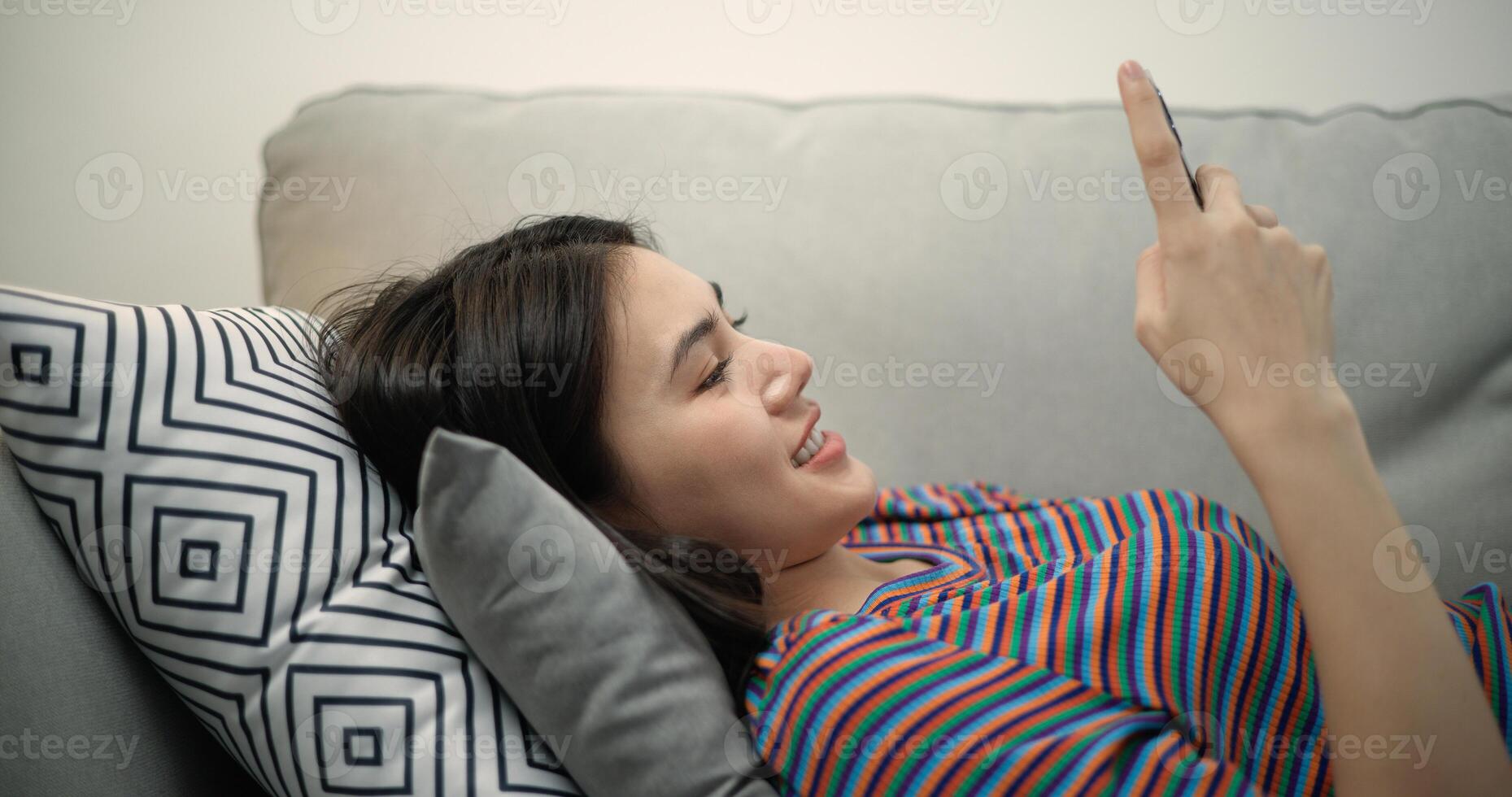 jong Aziatisch vrouw glimlachen en surfing de internet Aan een mobiel telefoon terwijl aan het liegen Aan de sofa foto