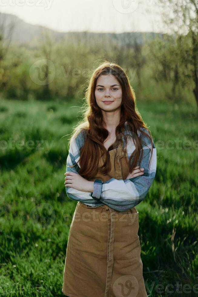 een boer vrouw in haar werk kleren, plaid overhemd en schort, staat in de veld- Aan de groen gras en glimlacht in de instelling zon foto