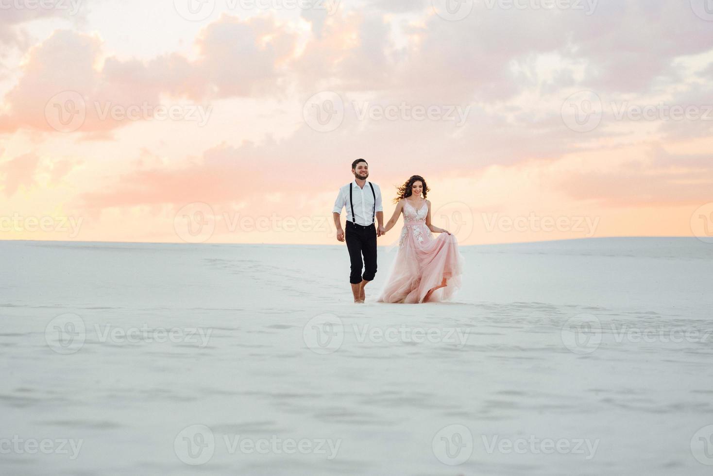 jong stel een man in zwarte rijbroek en een meisje in een roze jurk lopen langs het witte zand foto