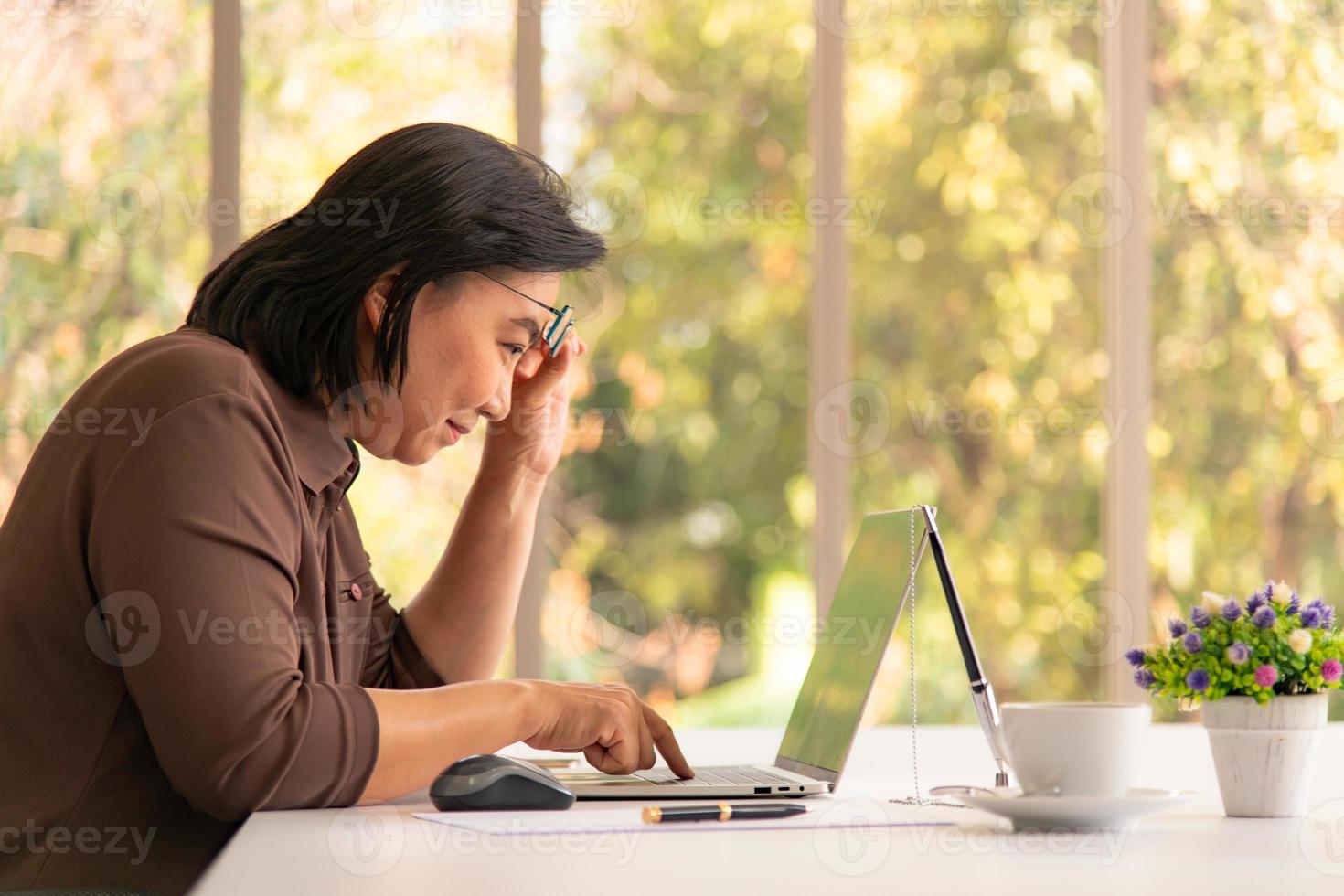 senior Aziatische vrouw die aan laptop op de natuurlijke woonkamerachtergrond werkt met moderne technologie foto