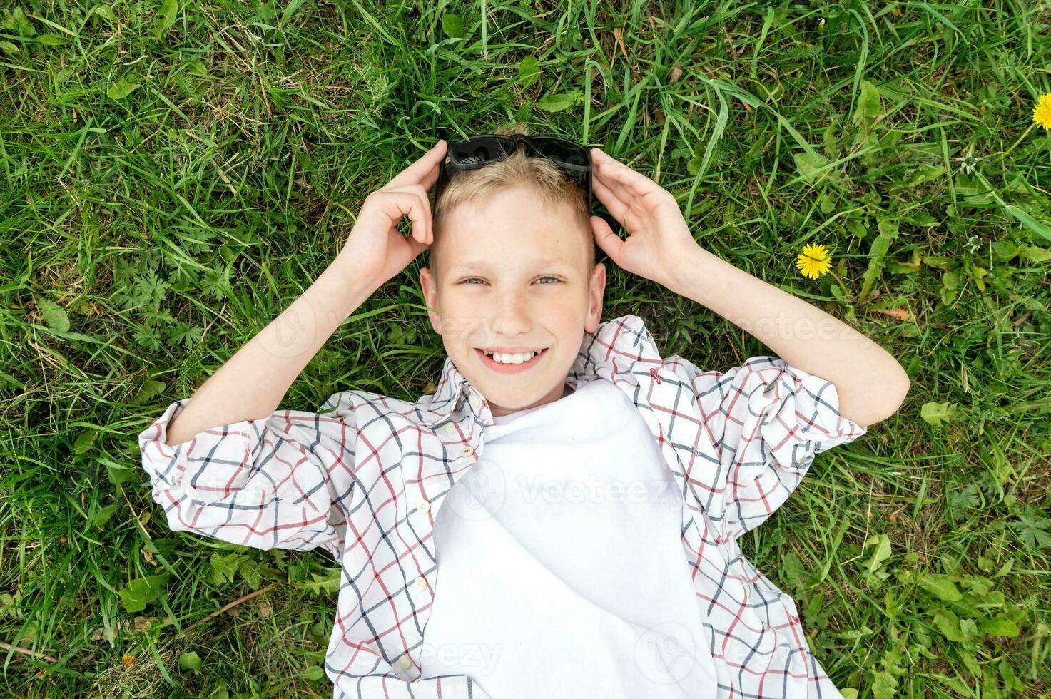 een schattig jongen leugens in de gras en glimlacht, houdt zonnebril Aan zijn hoofd foto