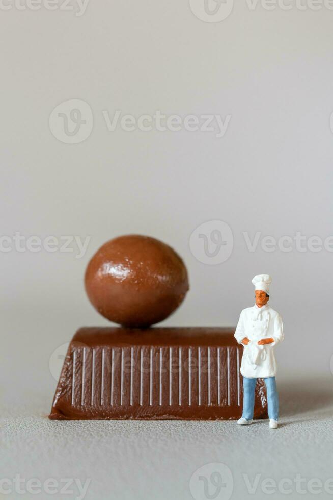 miniatuur mensen chef met chocola staand terwijl staand tegen een grijs achtergrond foto