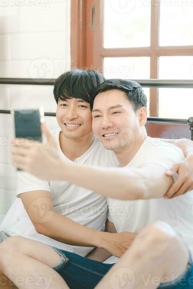 gelukkig Aziatisch homo paar gebruik mobiel telefoon video telefoontje of selfie Aan bed, lgbtq concept. gelukkig homo paar hebben pret gebruik makend van mobiel smartphone in bed. verscheidenheid van lgbt verhoudingen. foto