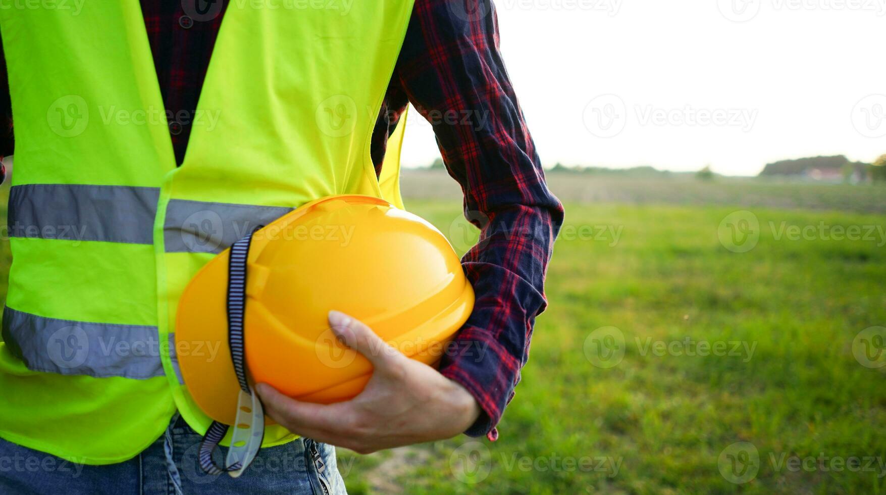 bouw arbeider Holding zijn geel helm, moeilijk hoed en bouw plan vervelend fluorescerend vest Aan de leeg veld. beroeps veiligheid en Gezondheid osh foto