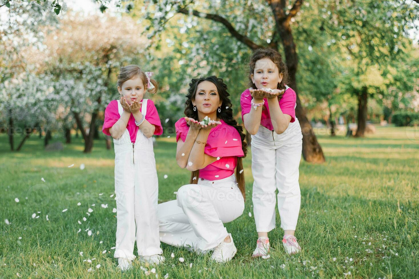 schattig mam en twee dochters blazen Aan de gedaald bloemblaadjes van appel bloesems in de voorjaar tuin. buitenshuis werkzaamheid. zoet momenten familie foto