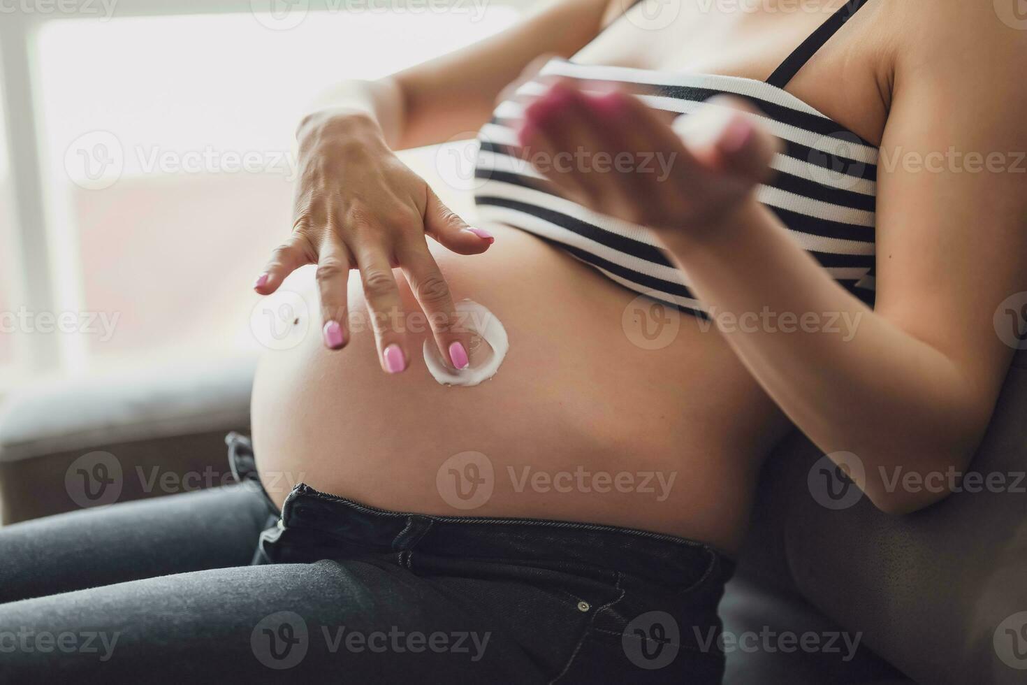 zwanger vrouw ontspannende Bij huis. ze is zittend Aan bed en toepassen huid room naar haar buik. foto