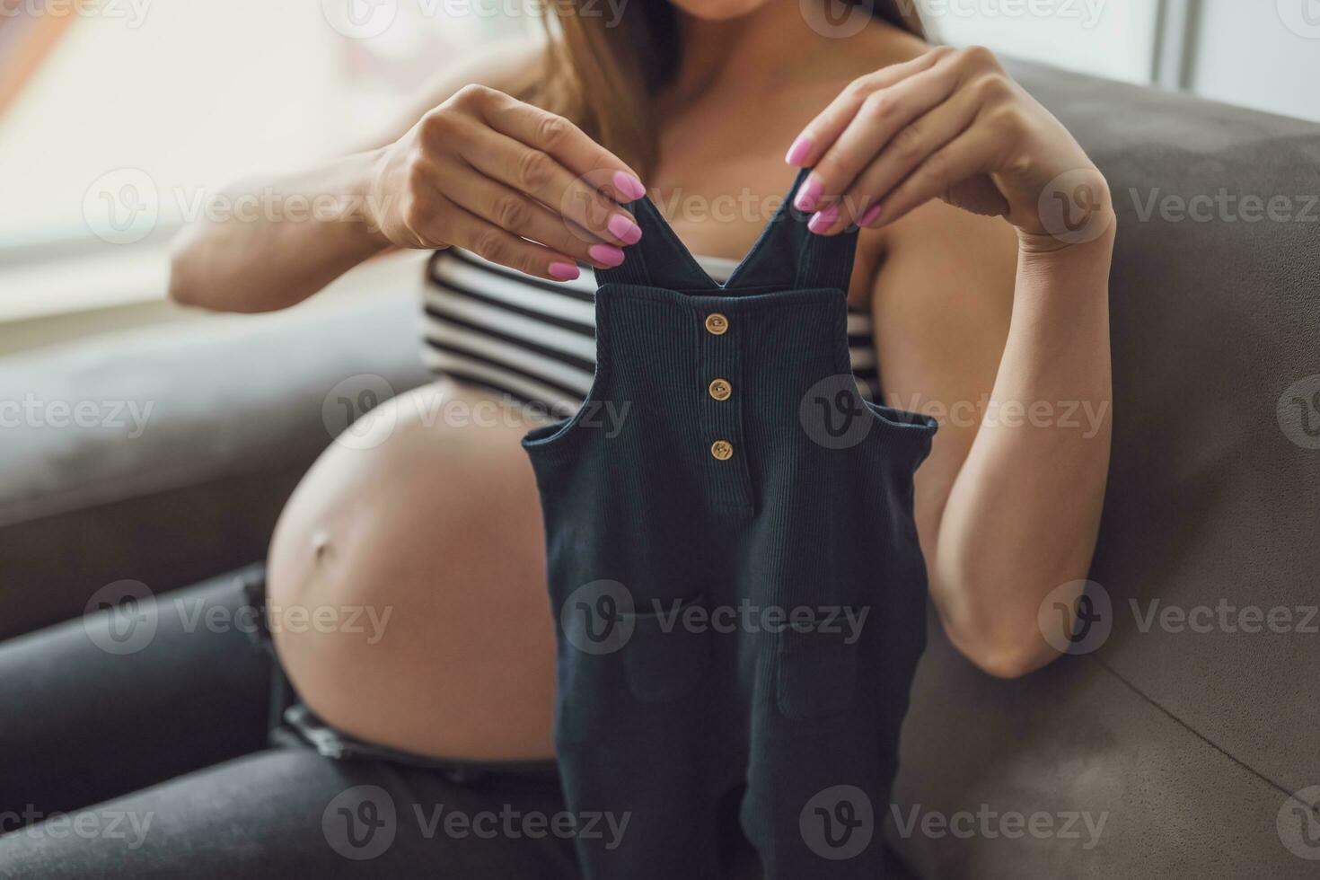 zwanger vrouw ontspannende Bij huis. ze is zittend Aan bed en Holding baby kleren. foto