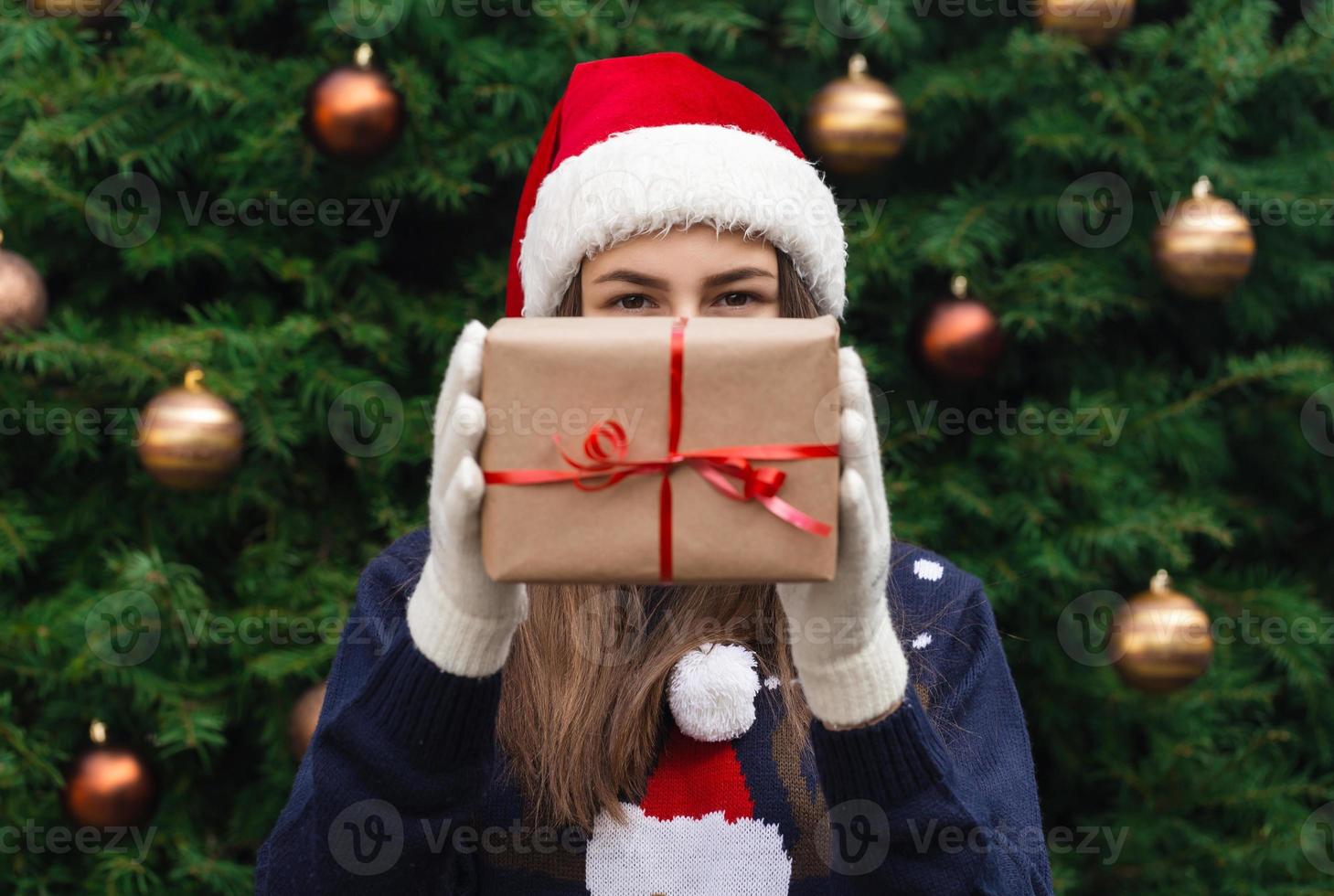 een jong meisje met een kerstmuts geeft een cadeau gemaakt van ambachtelijk papier met een rood lint foto