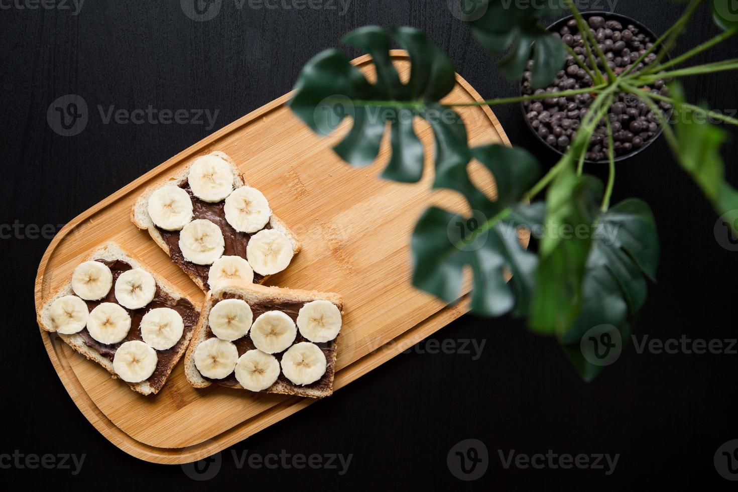 drie bananen wit brood toast verspreid met chocolade boter op een snijplank met bladeren op een donkere achtergrond foto