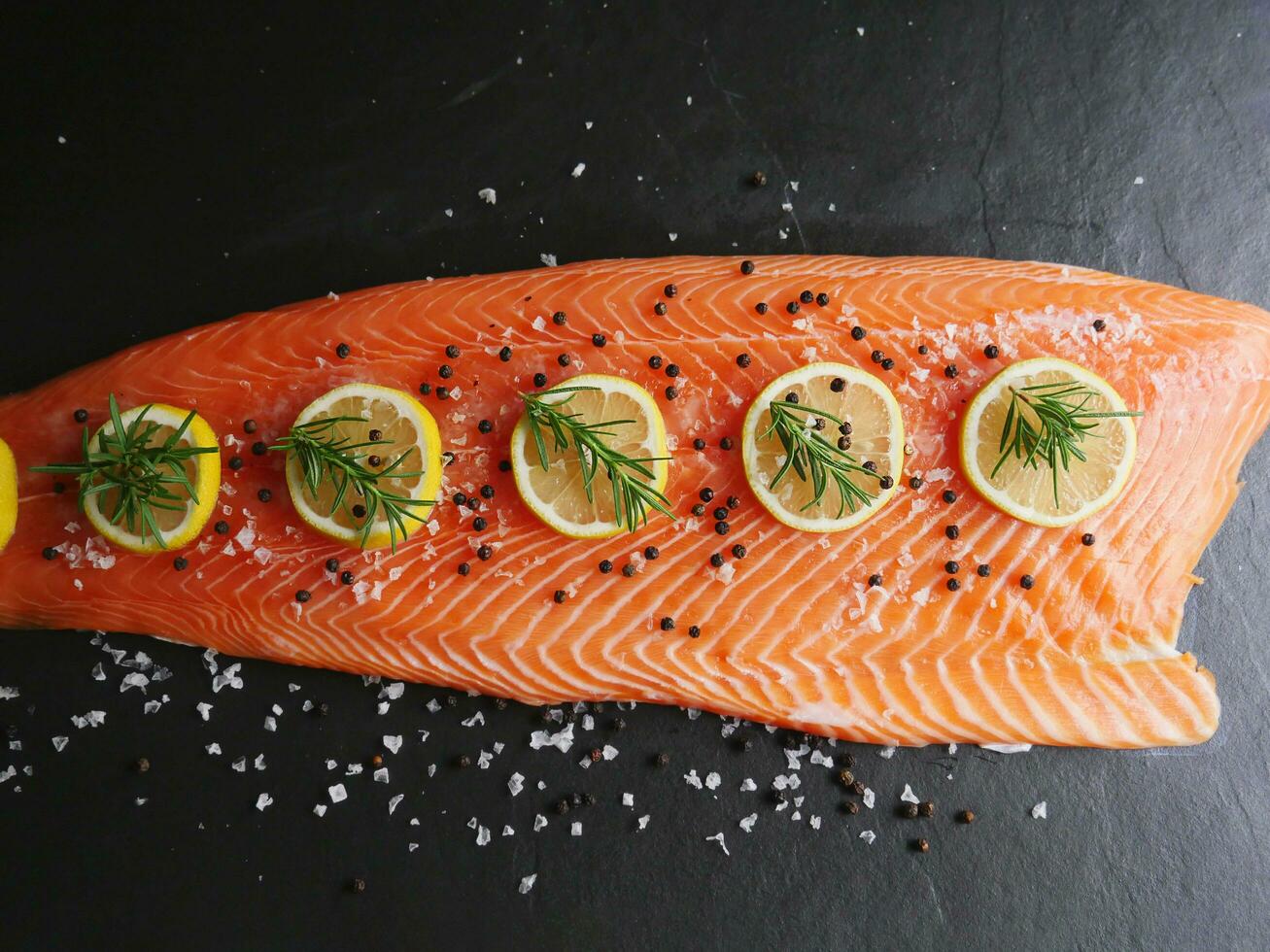 vers rauw Zalm forel filet met Koken ingrediënt kruiden rozemarijn peper en citroen Aan donker zwart steen oppervlak, voor de helft lichaam Zalm forel top visie, gezond rijk omega voeding voedsel voorafje foto