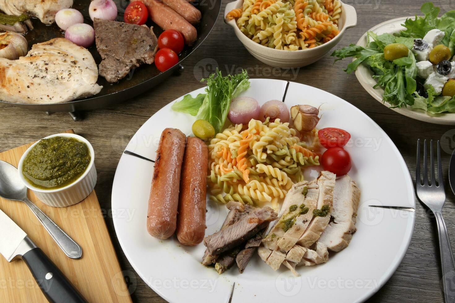 mengen ontmoeten kip rundvlees steak worst tomaat ui knoflook bakken rooster metaal pan kleurrijk pasta groen salade kaas olijf- munt basilicum pesto saus bord mes vork lepel gesneden geserveerd bord houten tafel foto