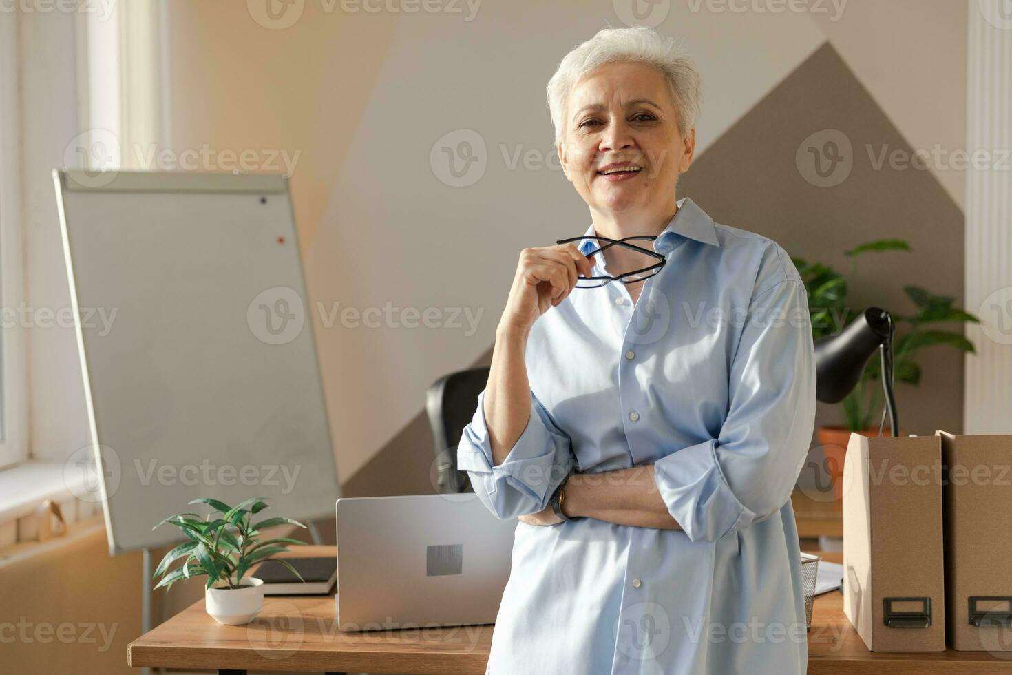 portret van zelfverzekerd elegant Europese midden- oud senior vrouw Bij werkplek. elegant ouder volwassen Jaren 60 grijs haren dame zakenvrouw in modern kantoor. baas leider leraar professioneel arbeider. foto