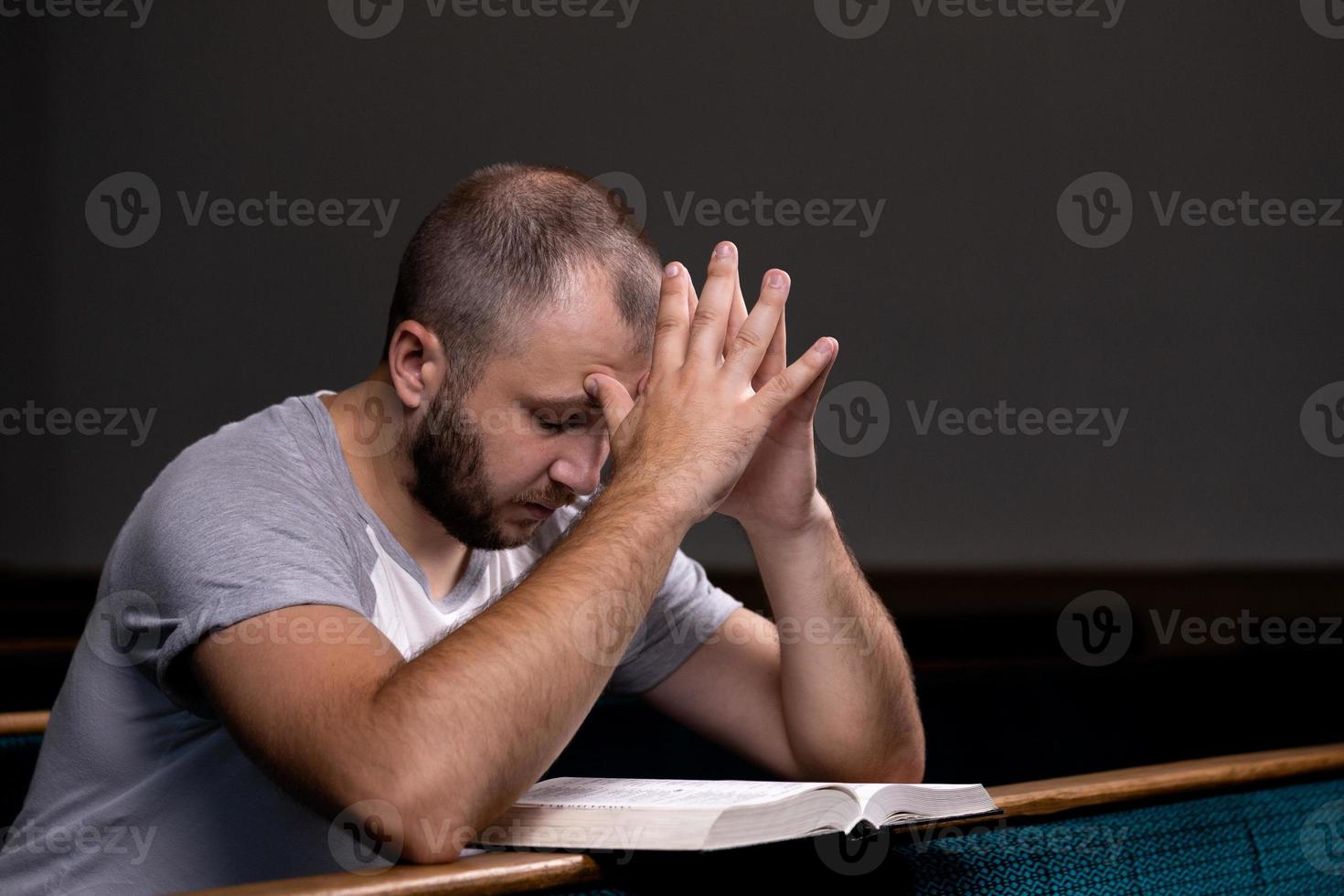 een jonge man zit op een kerkbankje, leest de bijbel en bidt foto