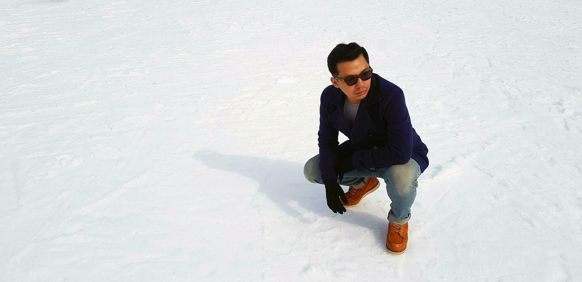 Aziatisch Mens in blauw jas of overjas en jeans, vervelend zonnebril, zitten Aan sneeuw achtergrond met kopiëren ruimte Bij Japan. toerist, ontspannend, knap, portret mode, mijlpaal voor reizen. verkoudheid temperatuur. foto