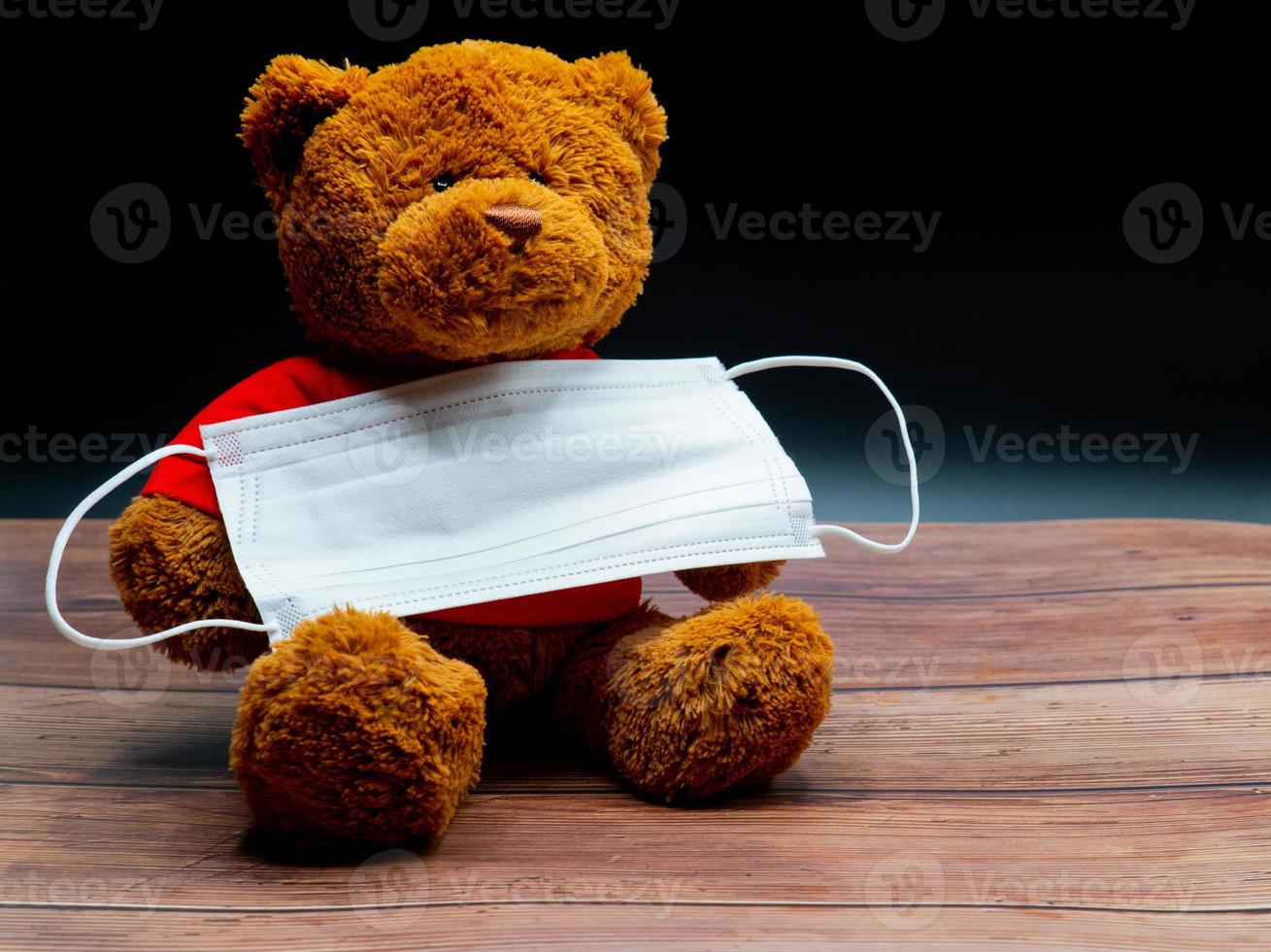 bruine teddybeer in rood t-shirt met een chirurgisch masker ter bescherming van coronavirus-infectie op de zwarte achtergrond foto