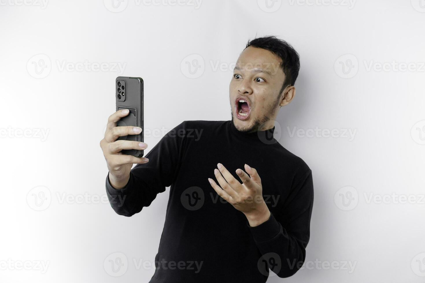 geschokt Aziatisch Mens vervelend zwart overhemd Holding zijn telefoon met zijn mond breed open, geïsoleerd door wit achtergrond foto
