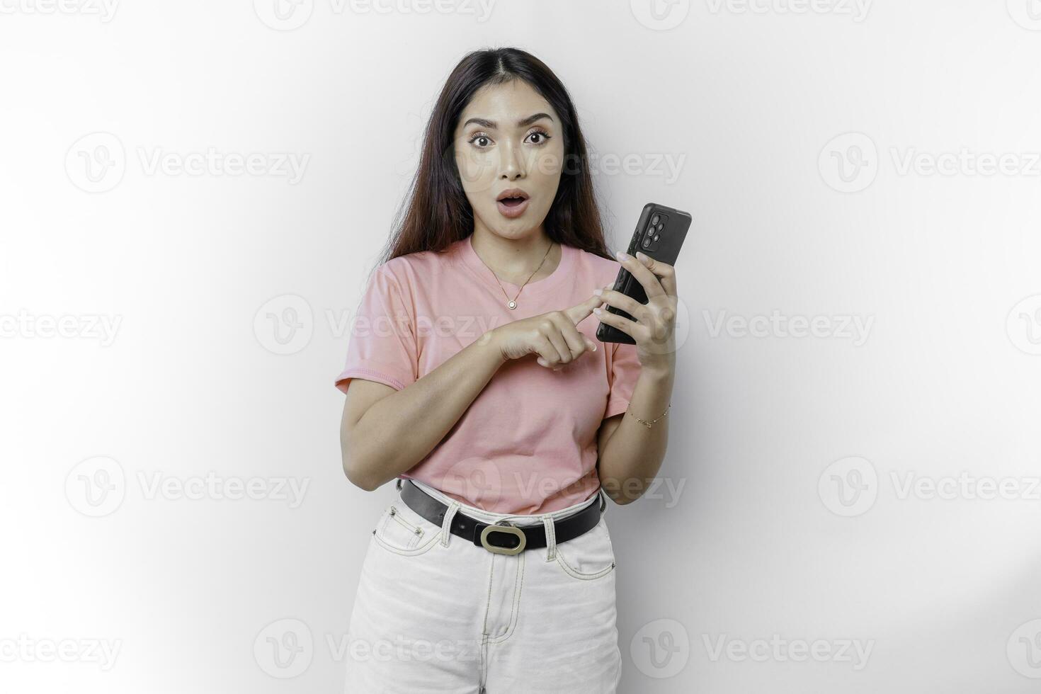 geschokt Aziatisch vrouw gekleed in roze en Holding haar telefoon, geïsoleerd door wit achtergrond foto