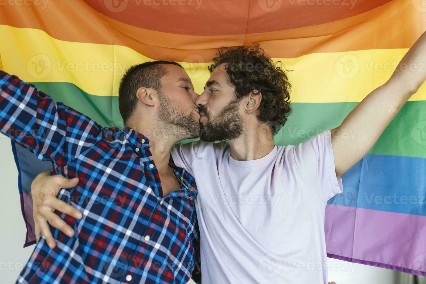 twee jong homo geliefden zoenen elk andere liefdevol. twee jong mannetje geliefden staand samen tegen een trots vlag. aanhankelijk jong homo paar sharing een romantisch moment samen. foto