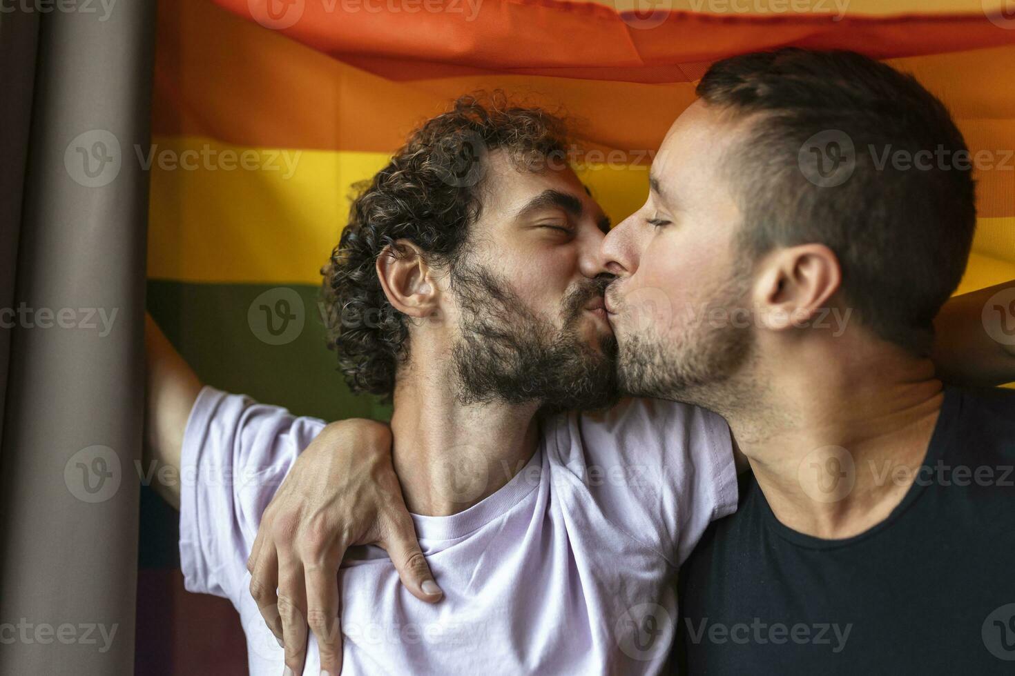 gepassioneerd homo paar maken uit met lgbtq vlag in de achtergrond. jong homo paar bonding liefdevol binnenshuis. foto