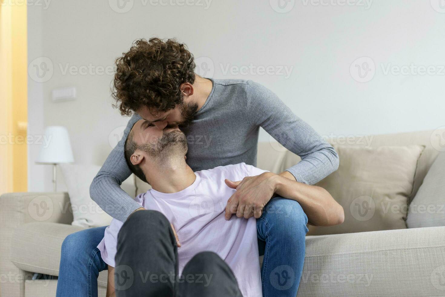 lief homo paar zoenen terwijl zittend in hun leven kamer Bij huis. twee romantisch jong mannetje geliefden hebben pret binnenshuis. jong homo paar leven samen. foto