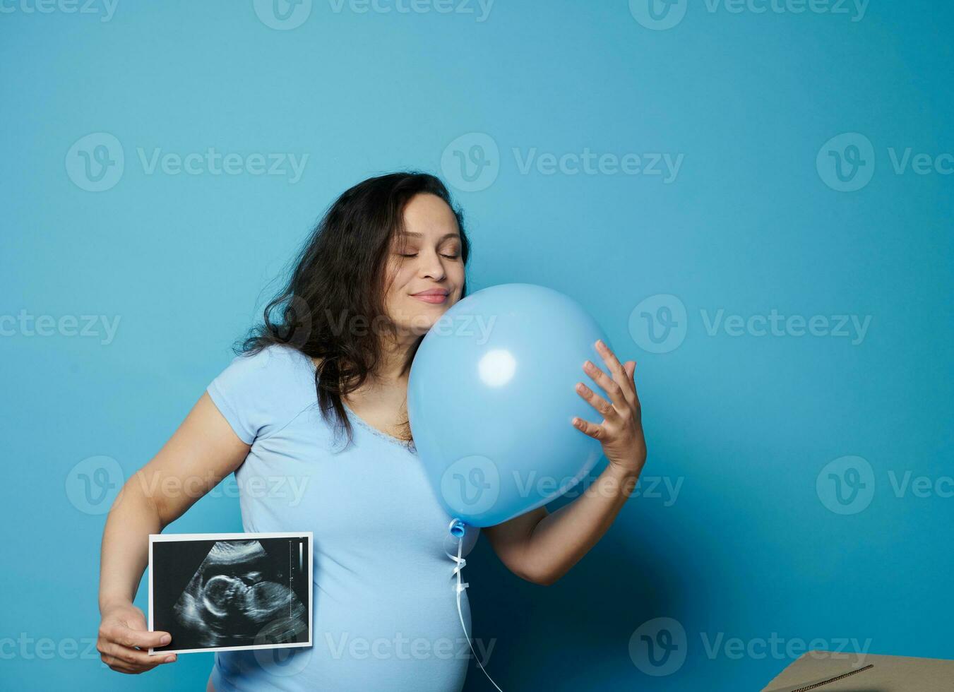 verrukkelijk zwanger vrouw knuffelen blauw ballon, poseren met echografie beeld van haar toekomst baby jongen, blauw backdrop foto