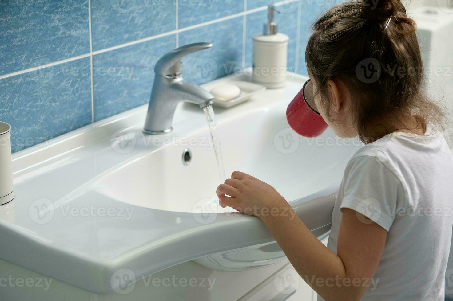 achterzijde visie aanbiddelijk kind meisje spoelen haar mond wanneer poetsen tanden, staand door wit keramisch wastafel in de huis badkamer foto
