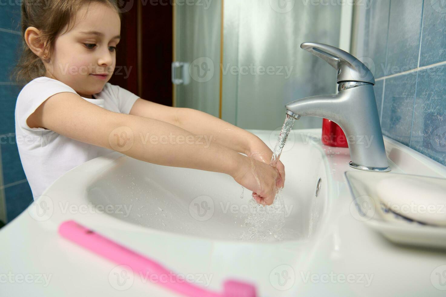 weinig kind meisje het wassen handen onder kraan met rennen water Bij huis badkamer. schoon handen, hygiëne en sanitair concept foto