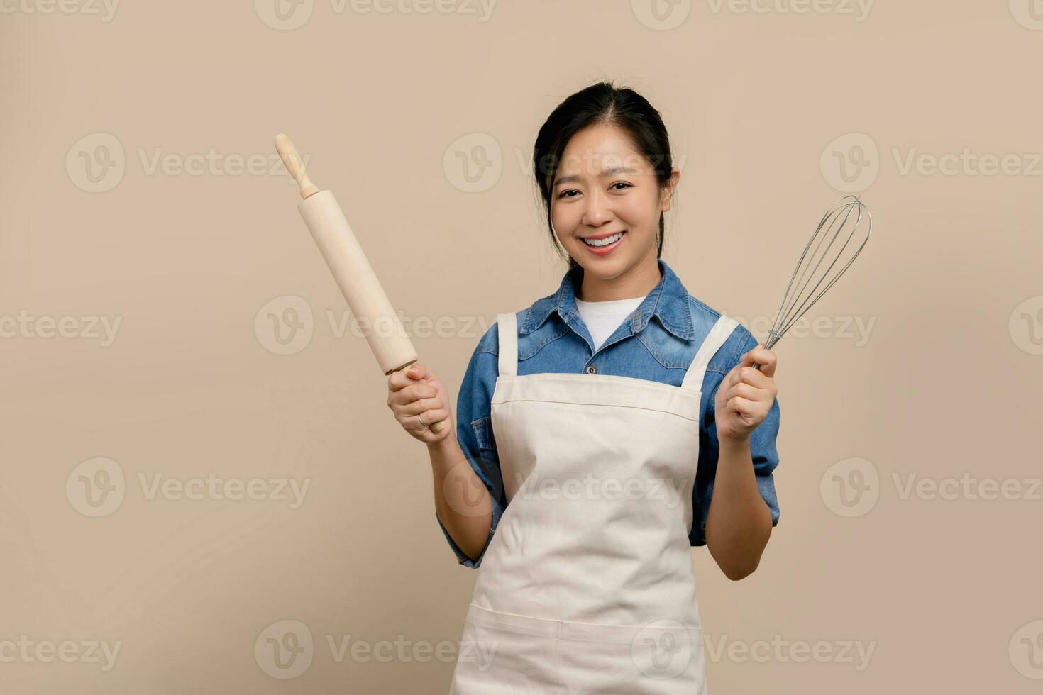 vrolijk Aziatisch vrouw vervelend schort en Holding bakken uitrusting in hand- staand geïsoleerd Aan licht bruin achtergrond. foto