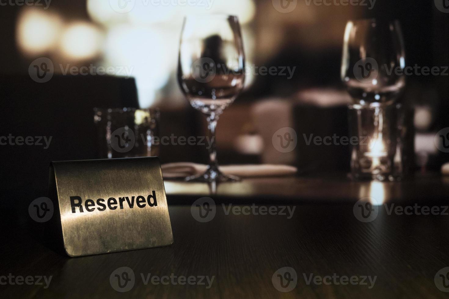 een metalen plaatje met de woorden gereserveerd staat links op een houten tafel in een restaurant met op de achtergrond een kaars en glazen foto