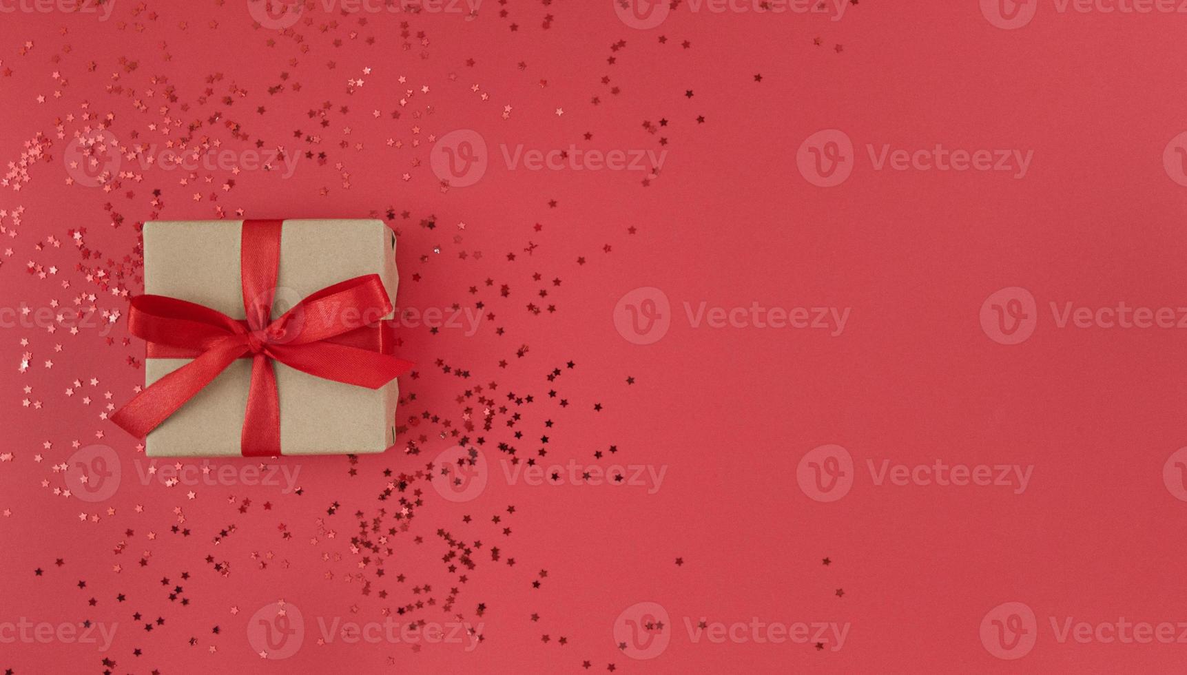 geschenkdoos verpakt in een ambachtelijk papier met rood lint met strik en confetti op rode achtergrond monochroom feestelijke plat leggen met kopie ruimte foto
