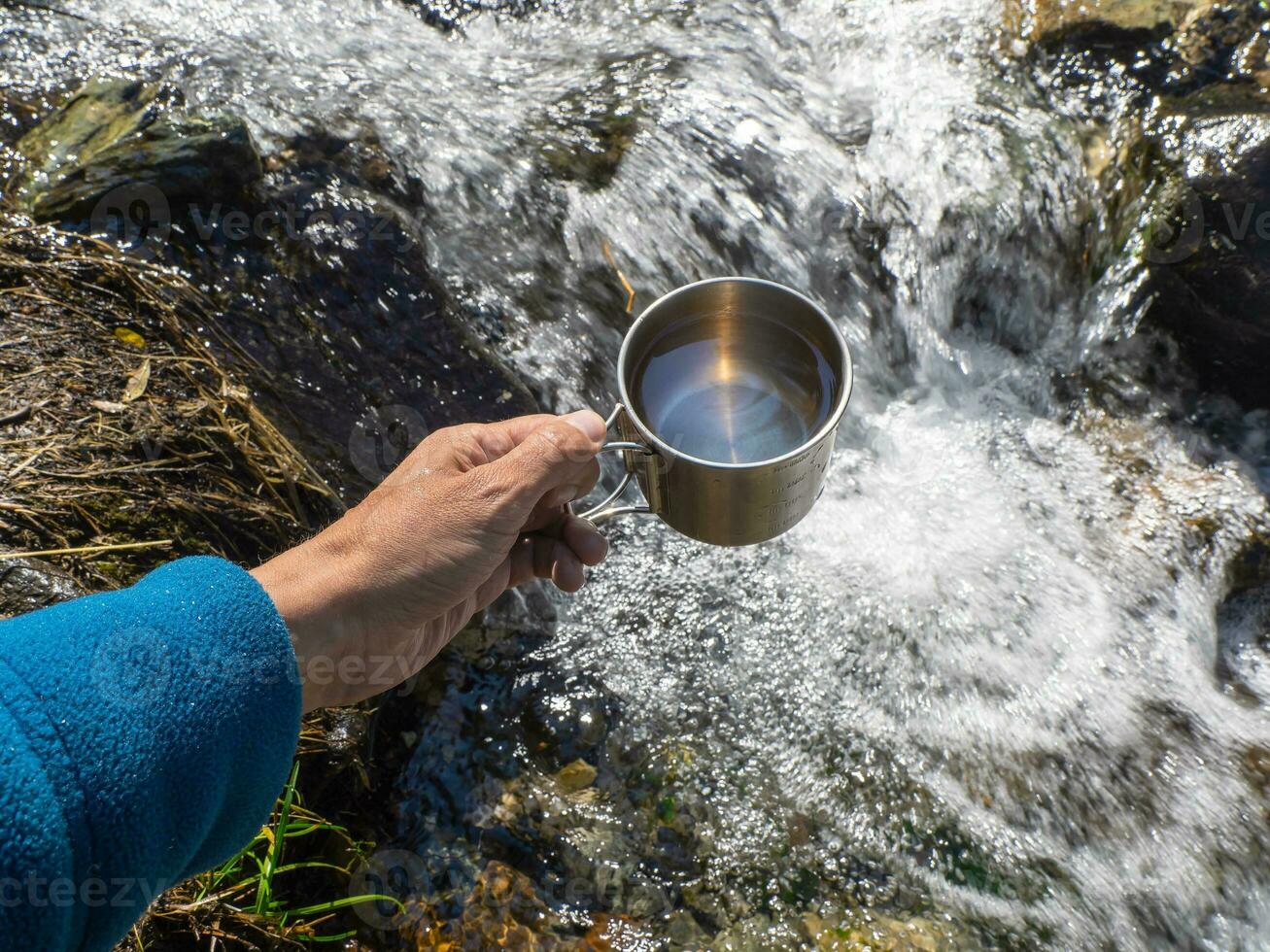 hand- Holding een wandelaars kop met schoon drinken water van een berg stroom. foto
