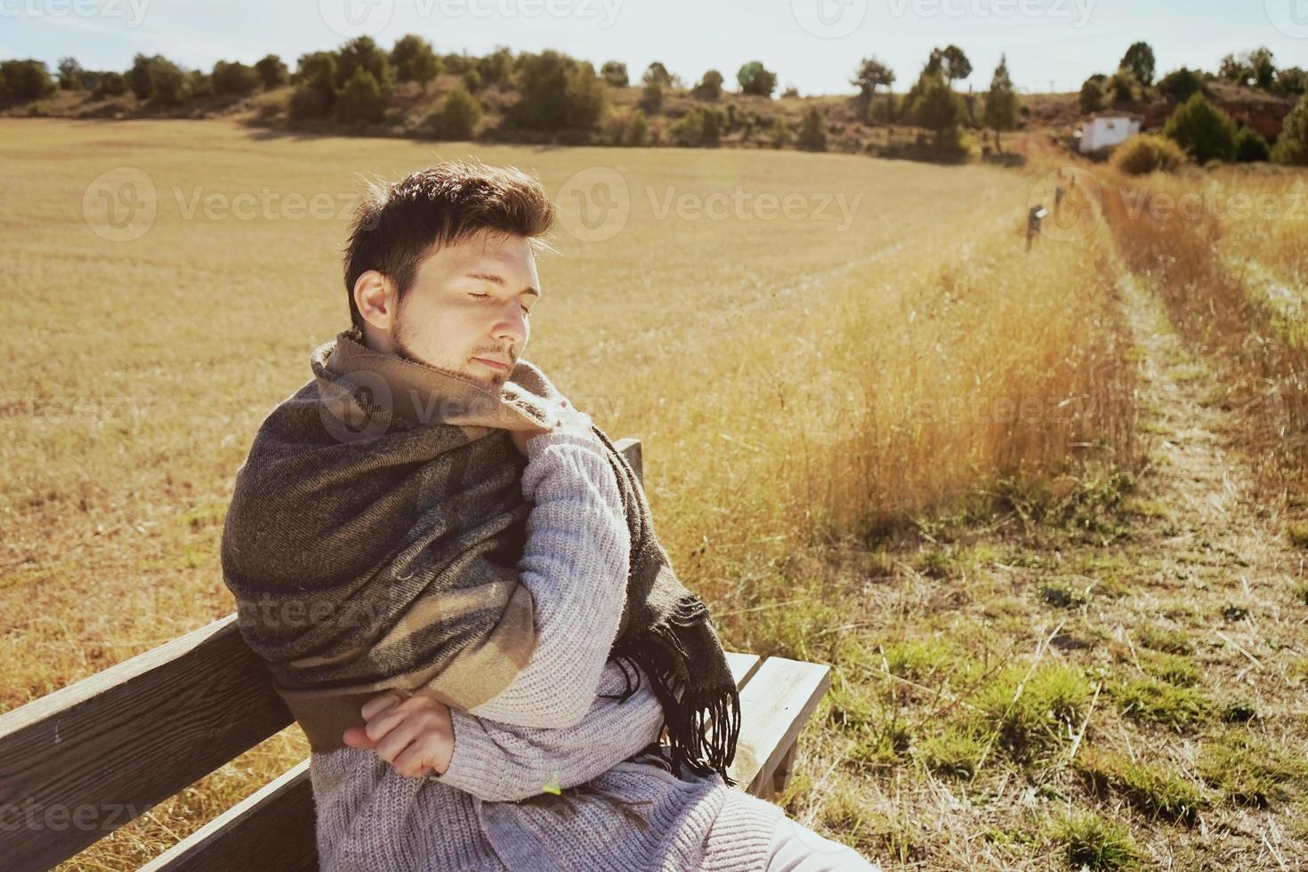 een jonge man met een warme sjaal die rustig geniet van de ochtendherfstzon in een geel veld met de achtergrondverlichting van de blauwe lucht foto