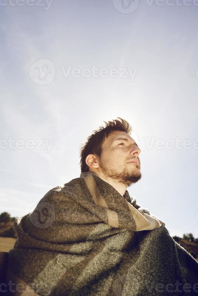 zijvlak van een jonge man met een warme sjaal die rustig geniet van de ochtendherfstzon met de achtergrondverlichting van de blauwe lucht foto