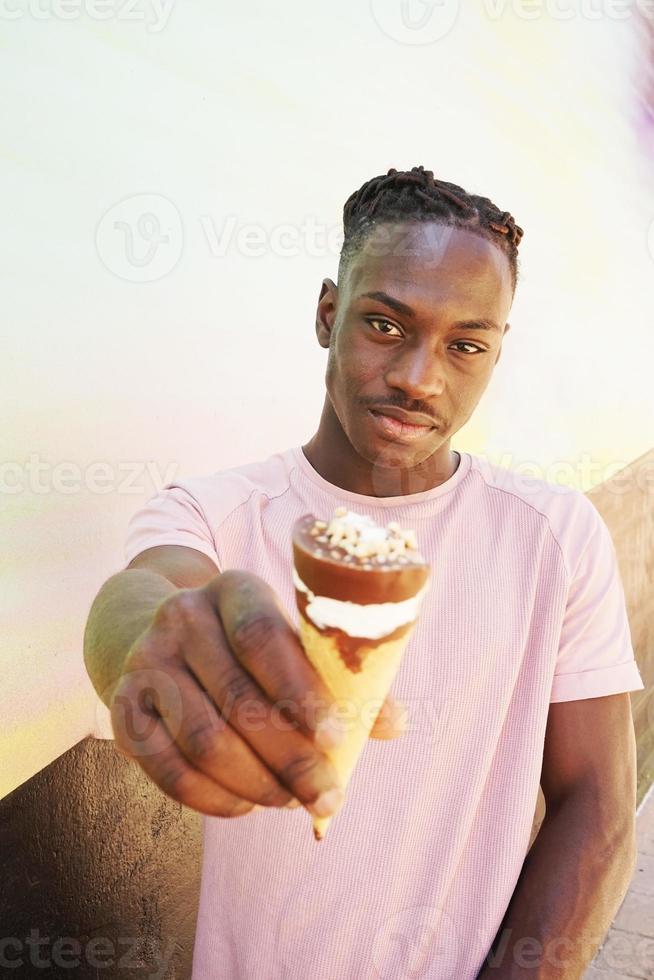 jonge knappe zwarte man draagt een roze shirt houdt en eet een ijshoorntje in de zomer op een geschilderde muur als een zonsopgang of zonnige dag foto