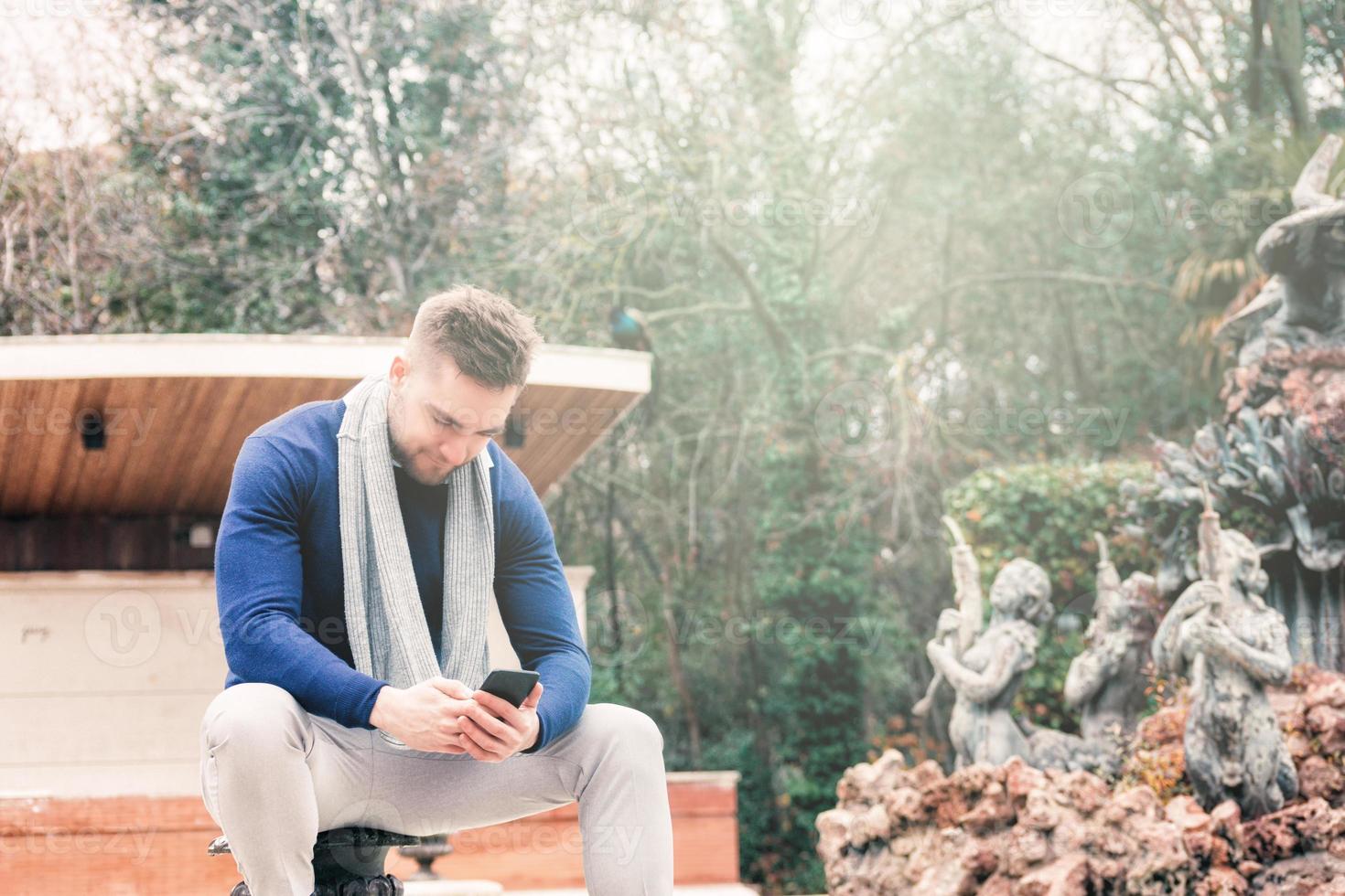 jonge man zit in de fontein van een park en kijkt naar zijn mobiele telefoon foto