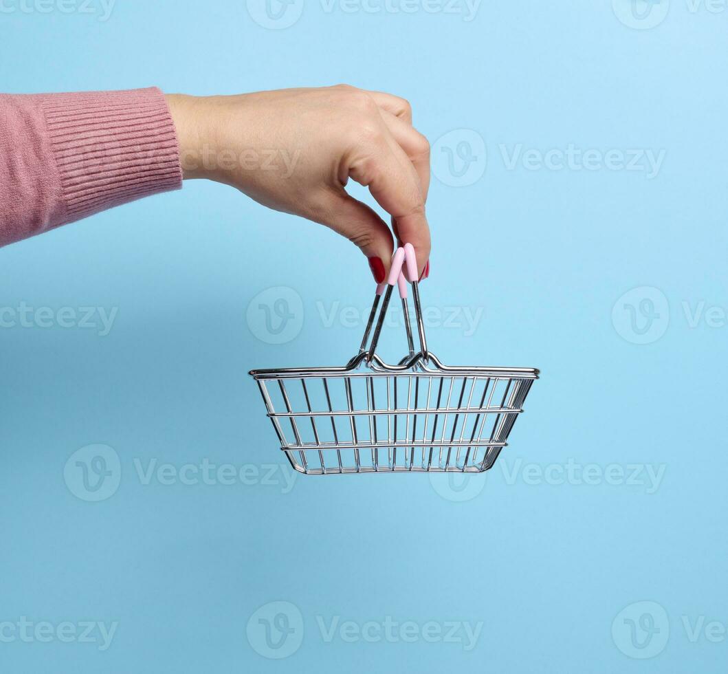 vrouw hand- Holding een miniatuur metaal boodschappen doen mand Aan een blauw achtergrond foto