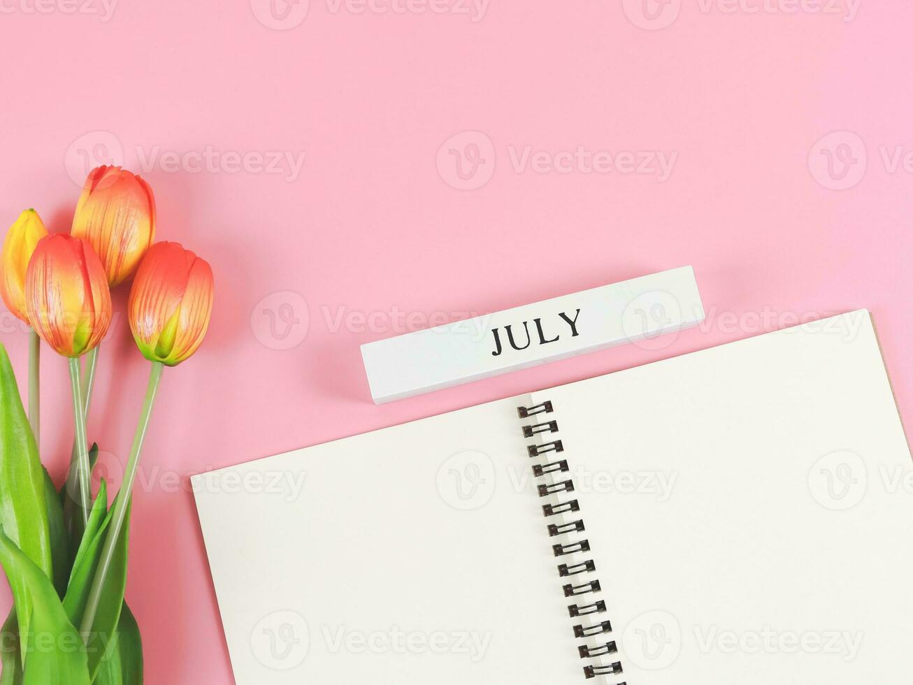 vlak leggen van geopend dagboek of notitieboekje met houten kalender juli Aan roze achtergrond met Oranje Geel tulpen. foto