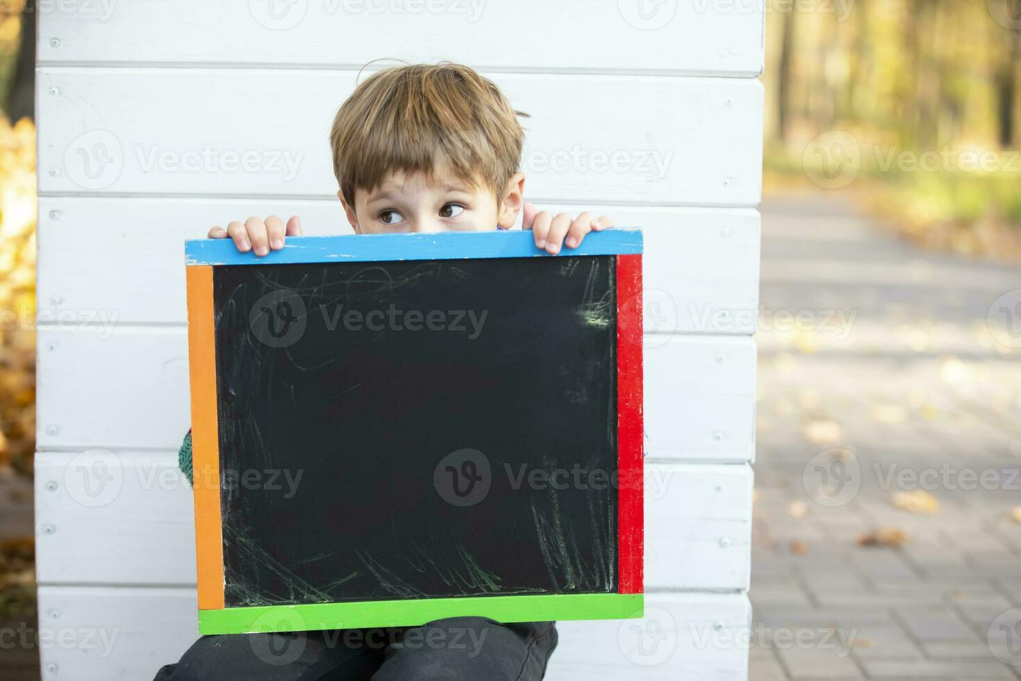 een weinig jongen zit in de park in de buurt de muur en houdt een leeg zwart bord voor de inscriptie. foto