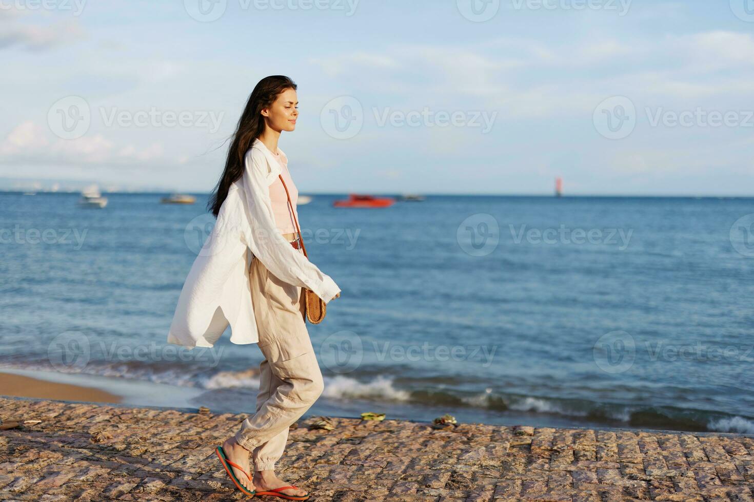 een vrouw gelukkig in de zon wandelen Aan de strand met haar armen verspreiding uit tegen de backdrop van de oceaan glimlach, vliegend haar, gebruind huid, ontspanning, reis naar de oceaan en vrijheid, zonsondergang licht Bali. foto