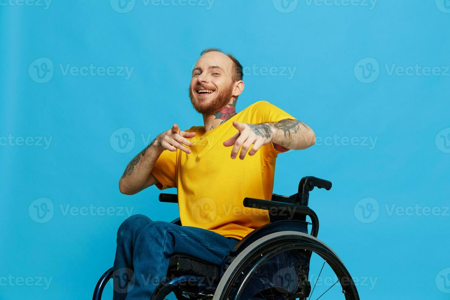 een Mens in een rolstoel glimlach looks Bij de camera in een t-shirt met tatoeages Aan zijn armen zit Aan een blauw studio achtergrond, een vol leven, een echt persoon foto