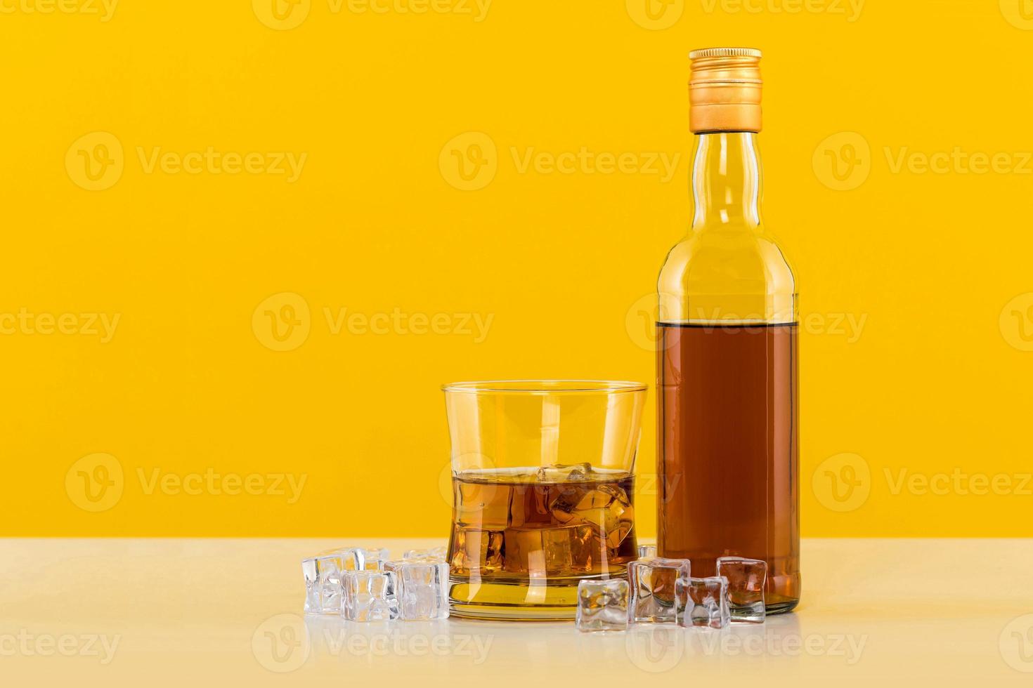 glas whisky met ijsblokjes en fles op gele achtergrond foto