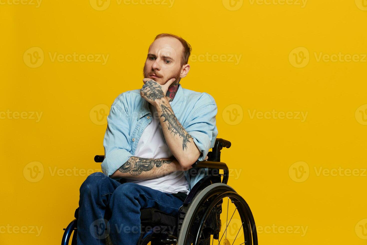 een Mens in een rolstoel problemen met de bewegingsapparaat systeem looks Bij de camera bedachtzaam met tatoeages Aan zijn armen zit Aan een geel studio achtergrond, Gezondheid concept een persoon met handicaps foto