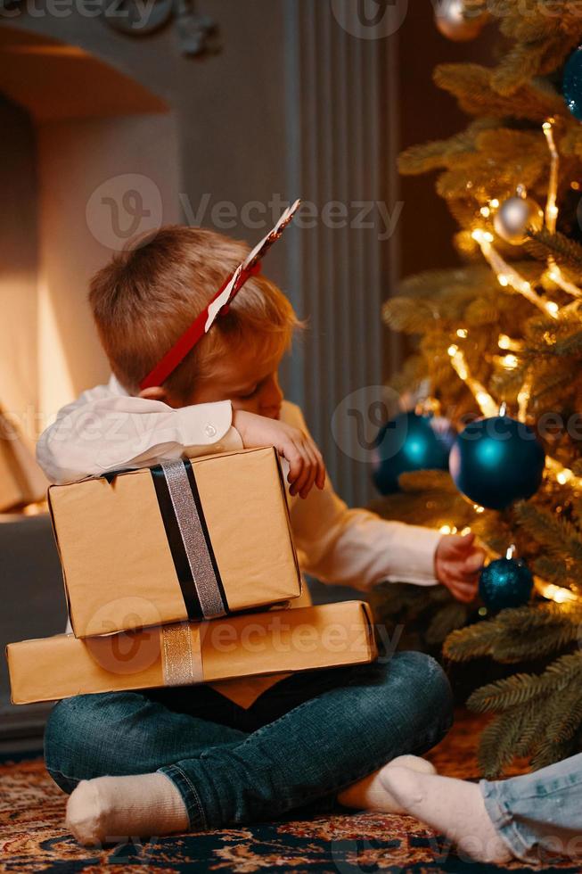 kleine jongen zit in de buurt van kerstboom met huidige dozen foto